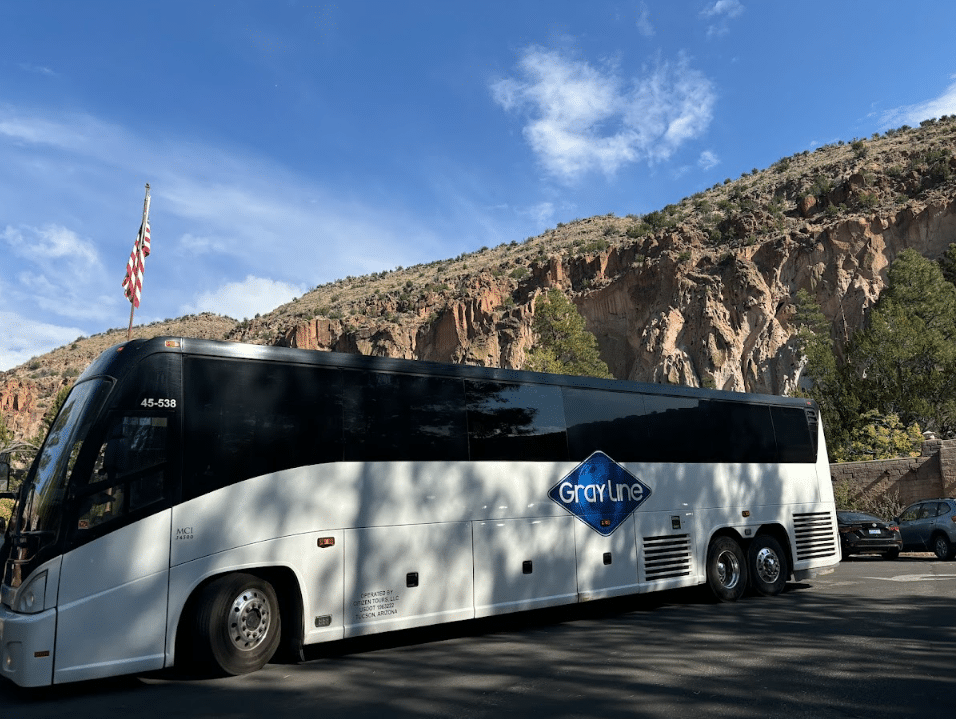The image shows a Gray Line charter bus in front of a mountainous backdrop, possibly near a scenic or historical site. Here's a suggested alt text: "Gray Line charter bus parked near rugged cliffs with a flag flying in the background, en route to a scenic destination.